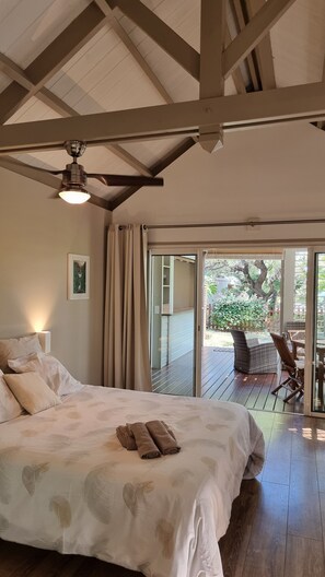 A bedroom with a bed, a ceiling fan, a view of an outdoor deck with chairs and a table, and a sliding glass door leading outside.