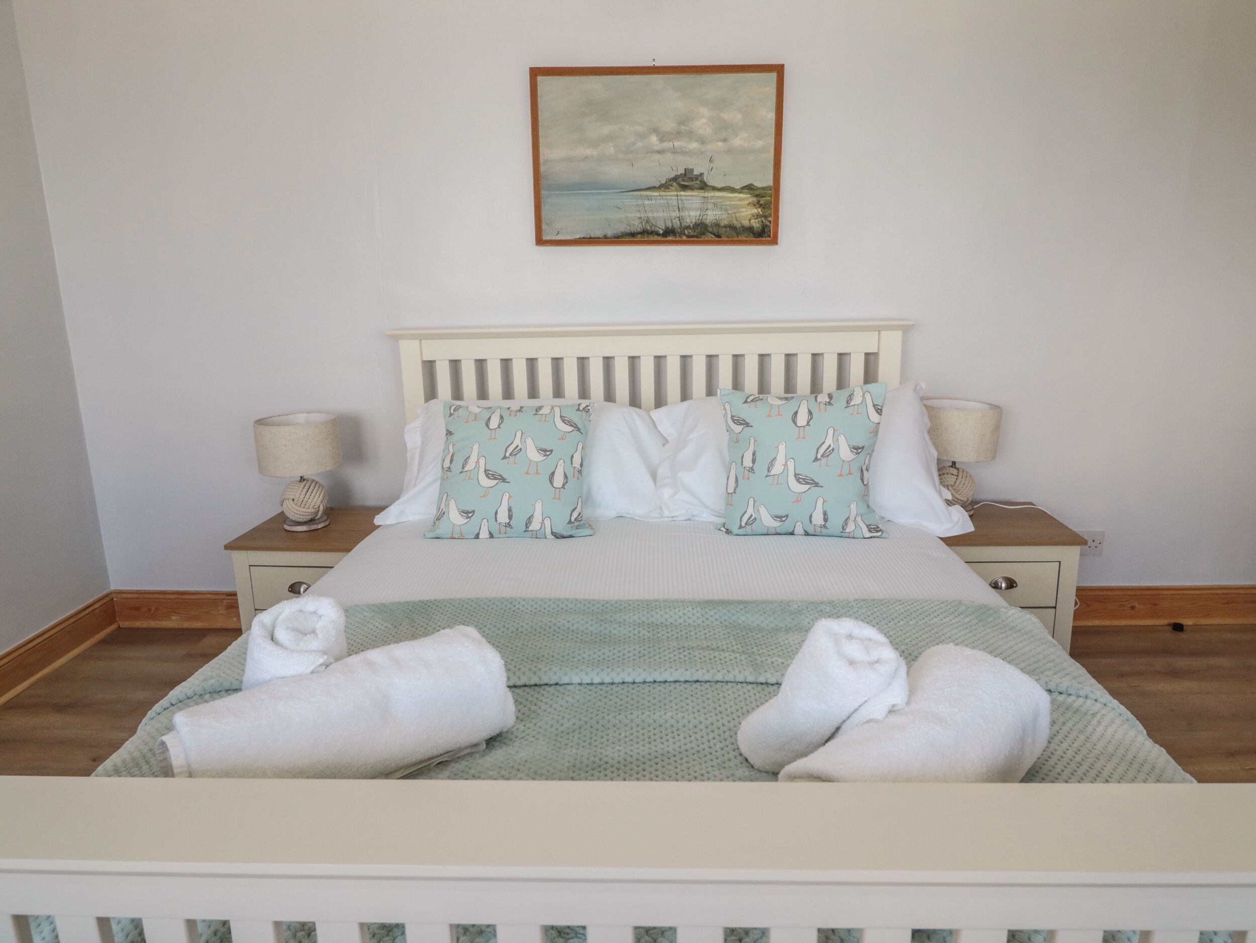 Een net opgemaakt bed met kussens en handdoeken, een schilderij erboven en nachtlampjes op houten nachtkastjes.
