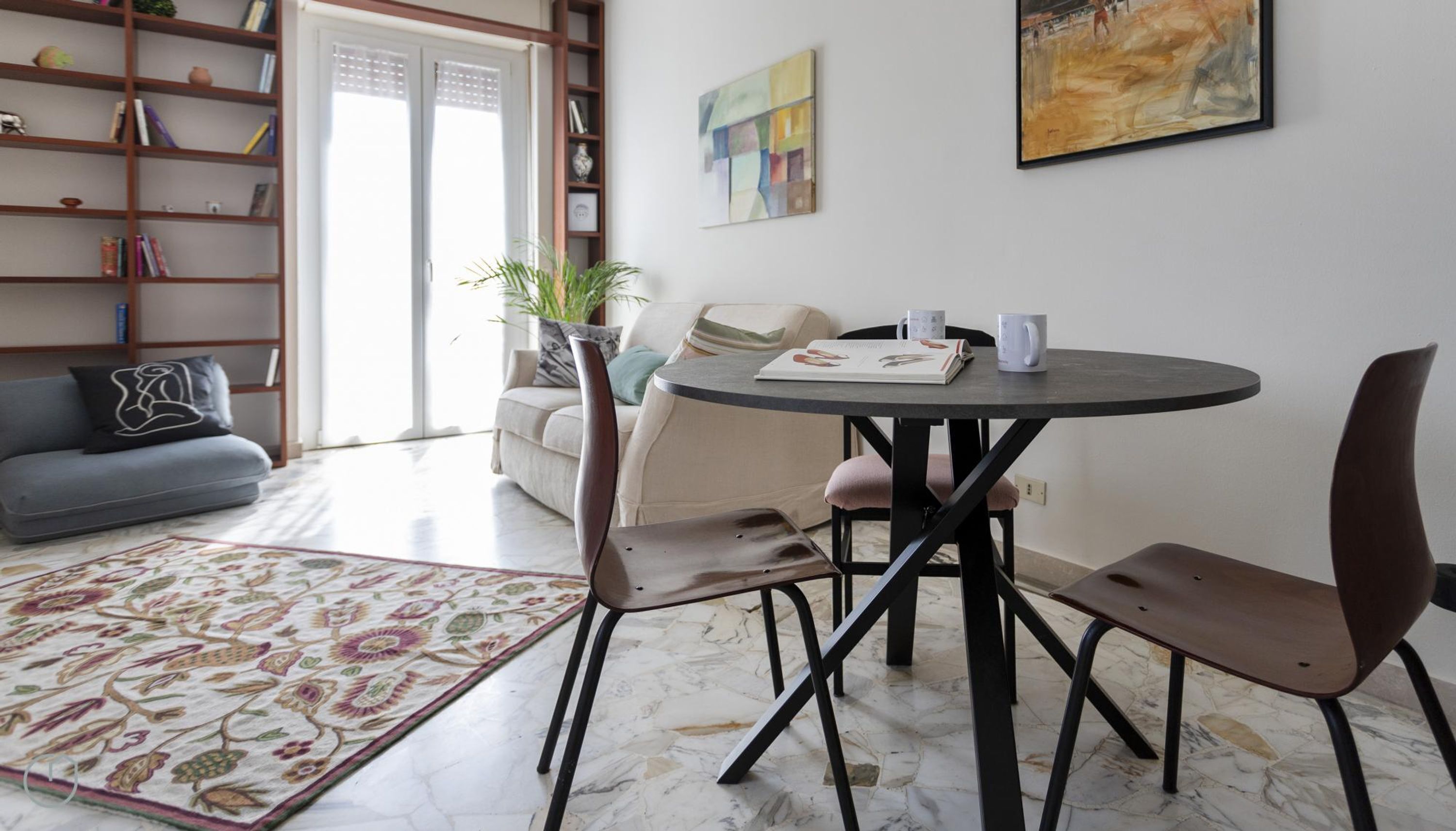 A modern living room with a round table, two chairs, a sofa, a bookshelf, and a painting on the wall.