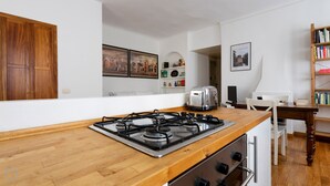 Une cuisine moderne équipée d’une cuisinière à gaz, d’un grille-pain et d’un plan de travail en bois. On y trouve une chaise blanche et une bibliothèque remplie de livres et d’objets décoratifs.