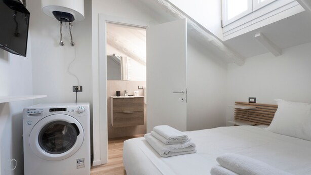 Ein modernes, gut beleuchtetes Schlafzimmer mit Waschmaschine, einem ordentlich bezogenen Bett, einer hölzernen Bank und einem großen Fenster.
