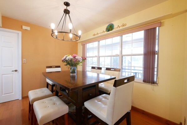A dining area with a wooden table, white cushioned chairs, a vase with flowers, and a window with curtains.
