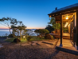 Una casa de madera con terraza, rodeada de árboles y vista al mar al atardecer.