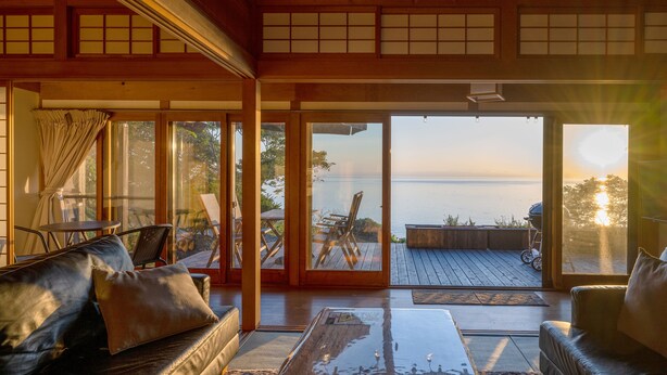 A spacious living room with a large sliding glass door leading to a deck overlooking a body of water. - Toya Cliff House (Toyako)