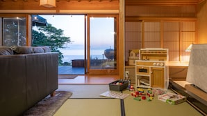 A traditional Japanese-style living room with tatami flooring, a sofa, a wooden cabinet with toys, and a view of the sea through large glass doors. - Toya Cliff House (Toyako)