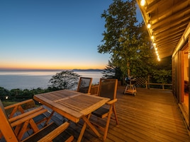 Una terraza de madera con mesa y sillas, con vistas a un cuerpo de agua y montañas al atardecer.