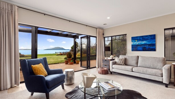 Uma sala de estar moderna com uma mesa de centro de vidro, uma poltrona azul, um sofá cinza e vista para o mar através de grandes portas de correr.
