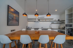 Een moderne keuken met een houten eettafel en stoelen, hangende lampen en uitzicht op zee door een groot raam.