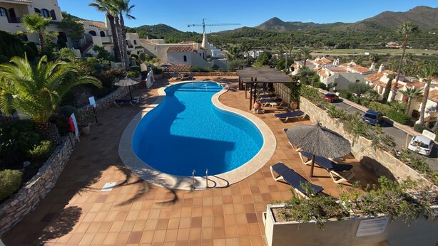 Una piscina curvada con sillas de descanso y sombrillas, rodeada de palmeras y montañas al fondo.