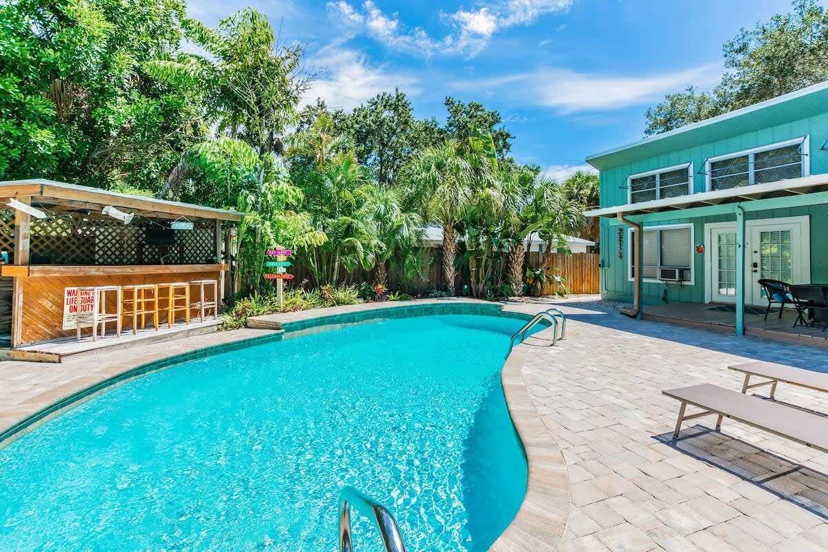 Une piscine dotée d’une terrasse en bois, entourée d’une végétation luxuriante et bordée d’un bâtiment à la façade de couleur turquoise.
