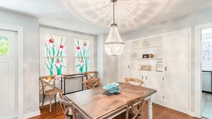 A dining area with a wooden table and chairs, a chandelier, and a window with floral artwork. - Downtown Dunedin B&B  Casa w/ Heated Pool (Dunedin)