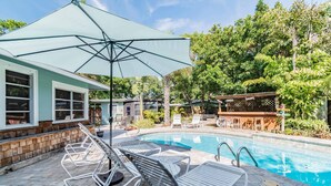 A poolside area with lounge chairs, a covered umbrella, and a wooden deck. - Downtown Dunedin B&B  Casa w/ Heated Pool (Dunedin)