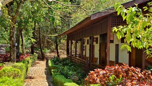 Un bâtiment traditionnel en bois, doté d’une allée couverte et entouré d’une végétation luxuriante, avec un chemin de pierre.
