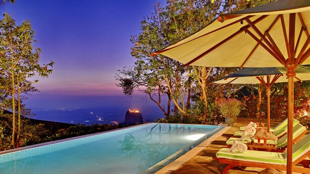 Une piscine entourée d’arbres, avec des chaises longues et des parasols, d’où l’on peut admirer une montagne lointaine au crépuscule.
