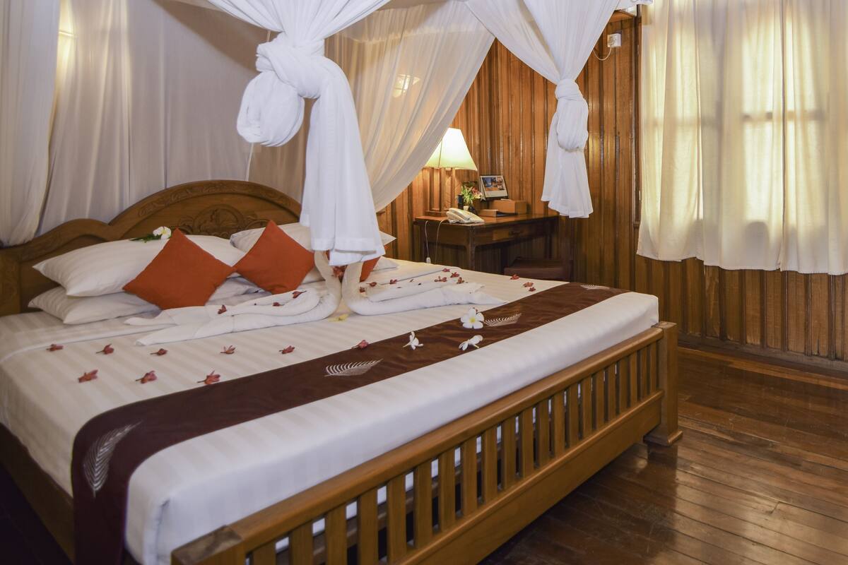 Cama de madeira com roupa de cama branca, almofadas laranja e manta marrom.