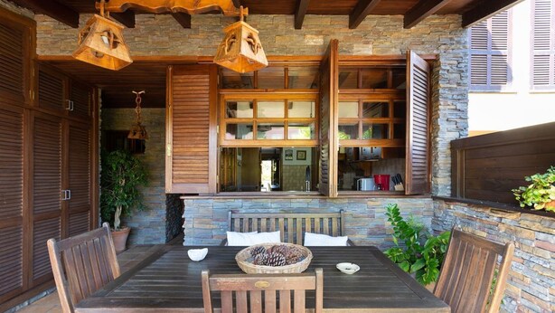 Un espace de restauration en plein air agrémenté d’un mur en pierre, de mobilier en bois et offrant une vue sur la cuisine grâce à de grandes fenêtres.