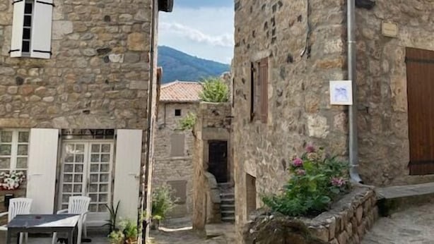 Un bâtiment en pierre doté d’un balcon, d’une porte en bois et d’un petit espace extérieur aménagé avec des chaises et des plantes.