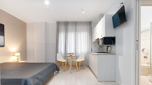 A compact hotel room with a bed, a small dining area, a kitchenette, and a TV mounted on the wall.