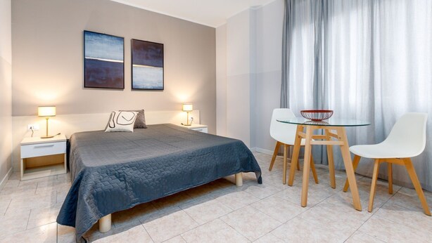 A modern bedroom with a large bed, bedside tables with lamps, a glass table with chairs, and artwork on the wall.