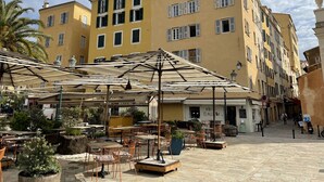 Un área de café al aire libre con mesas y sillas, una palmera y un edificio amarillo con ventanas y contraventanas.