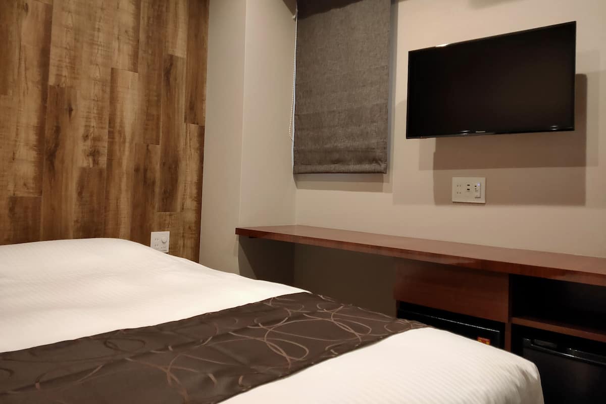 Quarto de hotel com cama, televisão fixada na parede, cabeceira de madeira e janela com persiana.