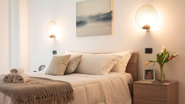 Ein ordentlich bezogenes Bett mit beige farbenem Duvet und Kissen, ein Nachttisch mit Blumen in einer Vase sowie Wandleuchten.