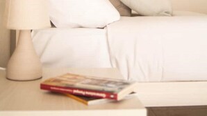 Ein ordentlich bezogenes Bett mit weißen Leinen und Kissen, eine Nachttischlampe und ein Buch auf dem Nachttisch.