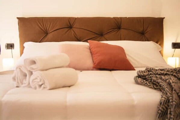 Una cama con cabecera capitonada, almohadas blancas y rojas, y toallas enrolladas.