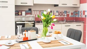 Eine moderne Küche mit weißen Schränken, einem Esstisch mit einer Tulpenvase, einer Kaffeemaschine und einem Teller Croissants.