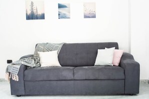 Un sofá gris con una manta y almohadas, frente a una pared lisa con tres cuadros enmarcados.