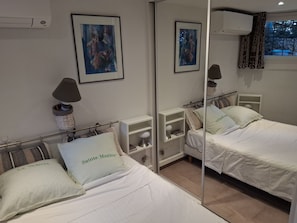 A hotel room with two beds, a mirror, a nightstand with a lamp, a framed painting, and a window with curtains.