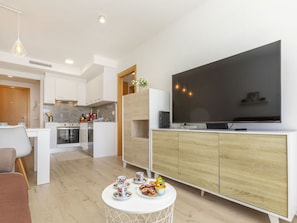 Uma sala de estar moderna com uma televisão de tela plana, uma área de jantar com mesa posta para dois e uma cozinha ao fundo.