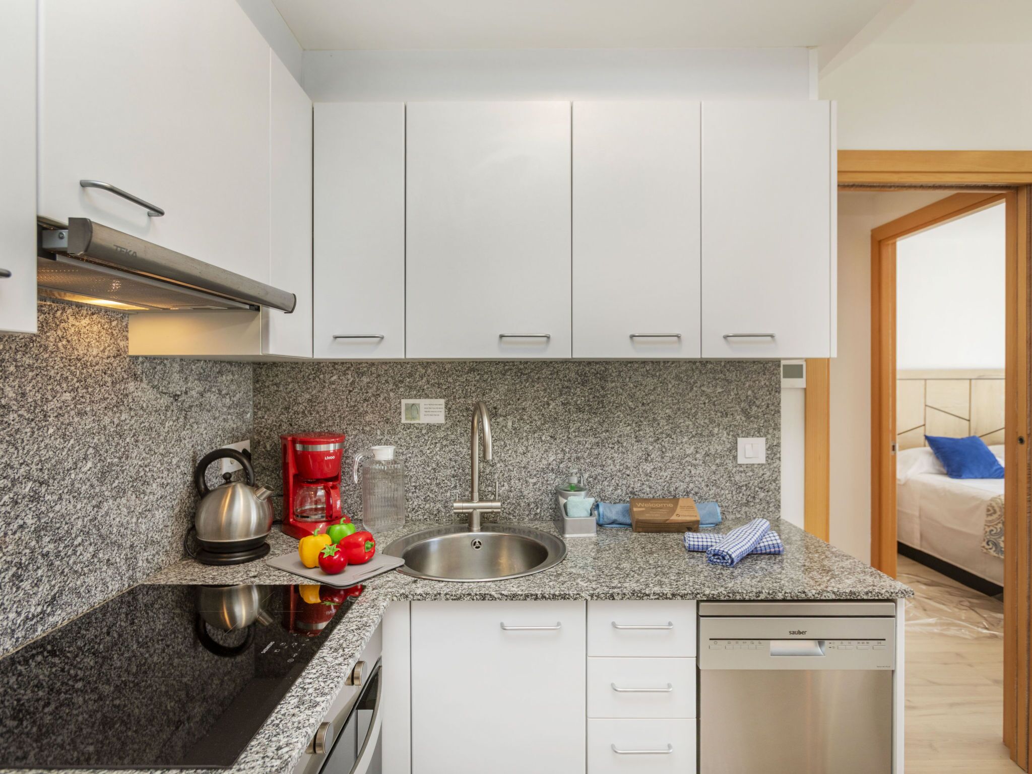 A modern kitchen with granite countertops, white cabinets, a coffee maker, a kettle, and a dishwasher.