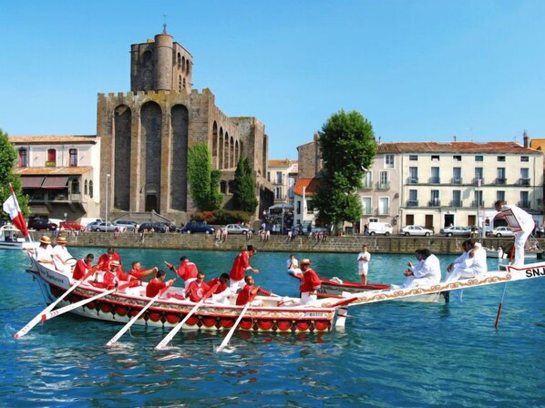 A traditional boat with rowers in red uniforms, a tower in the background, and buildings along the waterfront. - Primavera by Interhome (Cap d'Agde)