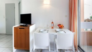 Ein modernes Zimmer mit Esstisch, zwei weißen Stühlen, einem Fernseher auf einer Holzkommode, einer Weinflasche, einem Glas und einer Obstschale.