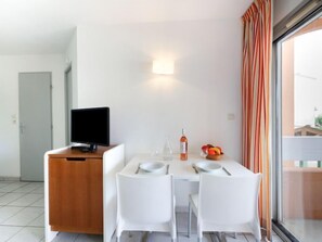 A modern room with a dining table, two white chairs, a television on a wooden cabinet, a bottle of wine, a glass, and a bowl of fruit. - Primavera by Interhome (Cap d'Agde)