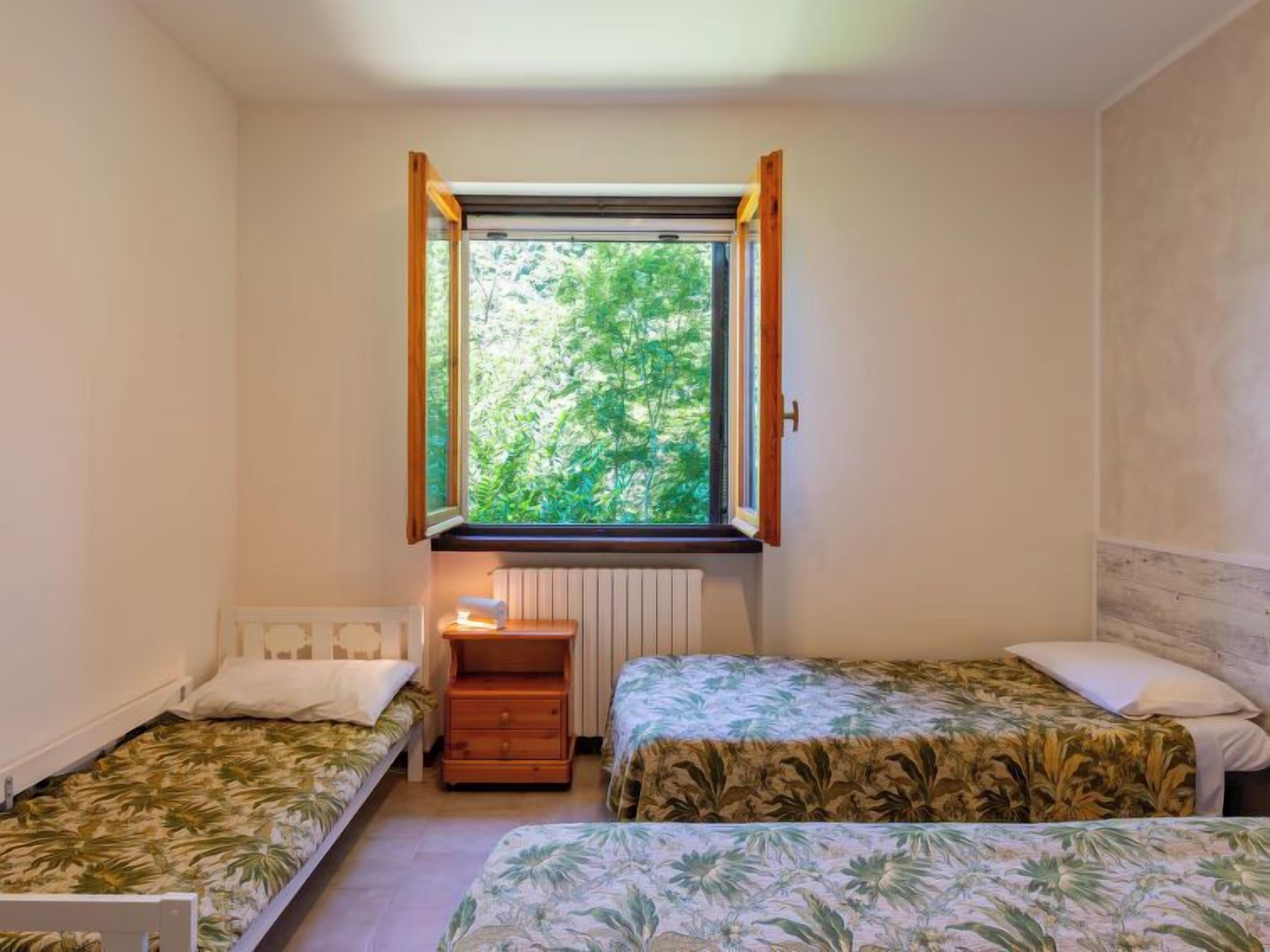 Una habitación pequeña y acogedora con dos camas individuales, una mesita de noche, un ventanal con vistas a la vegetación y un radiador.