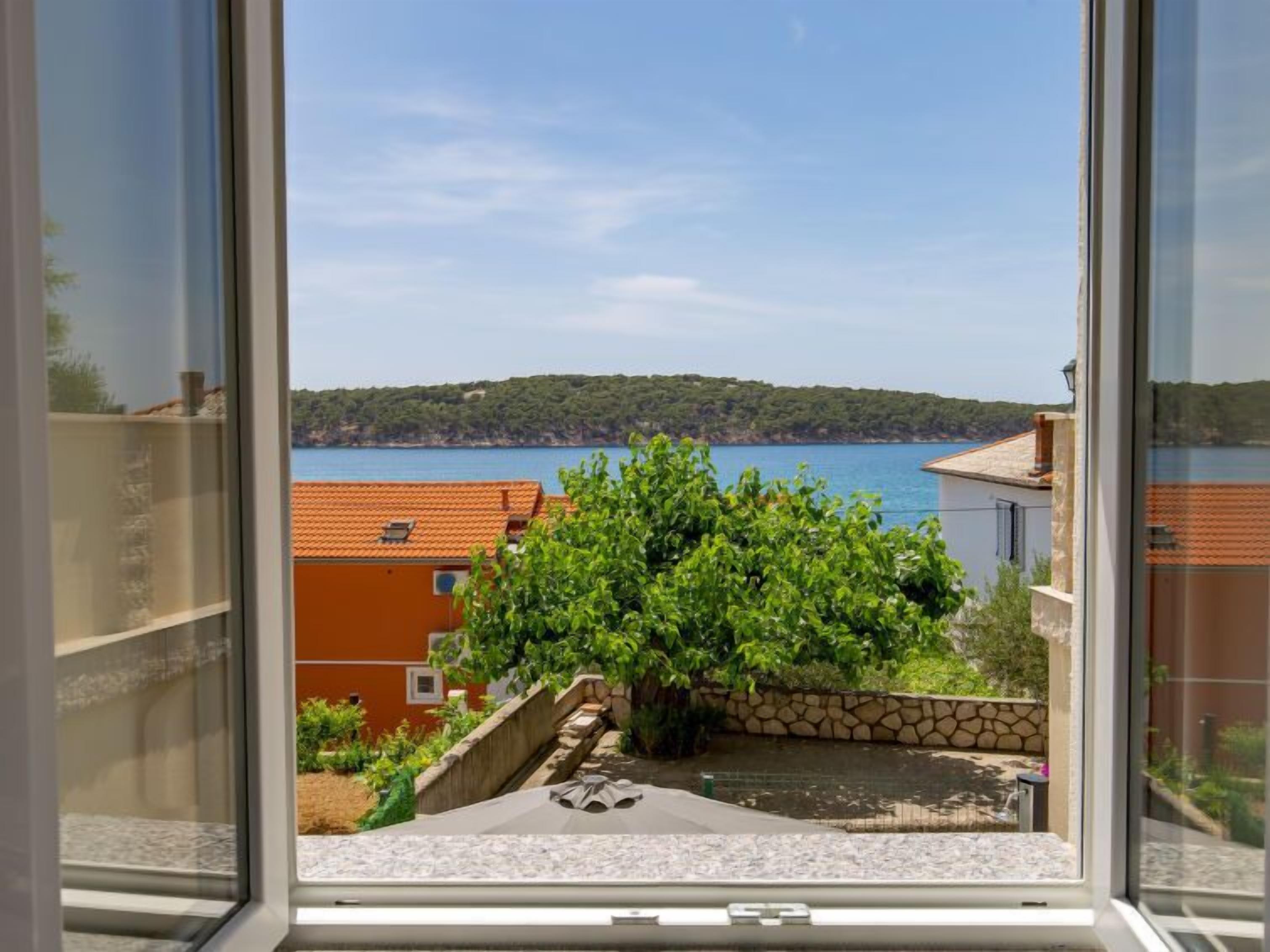 Blick aus einem Fenster auf einen Innenhof mit einer Steinmauer, einem kleinen Pool und im Hintergrund Meer und Hügel.