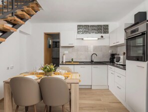 Uma cozinha moderna com armários brancos, uma mesa de jantar de madeira, duas cadeiras, forno embutido e uma escada.