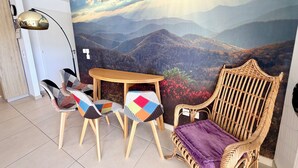A room with a mountain landscape mural, a round table, two chairs with colorful cushions, a wicker chair with a purple cushion, and a floor lamp. - Comfortable T3 with terrace and garage! (Aix-les-Bains)