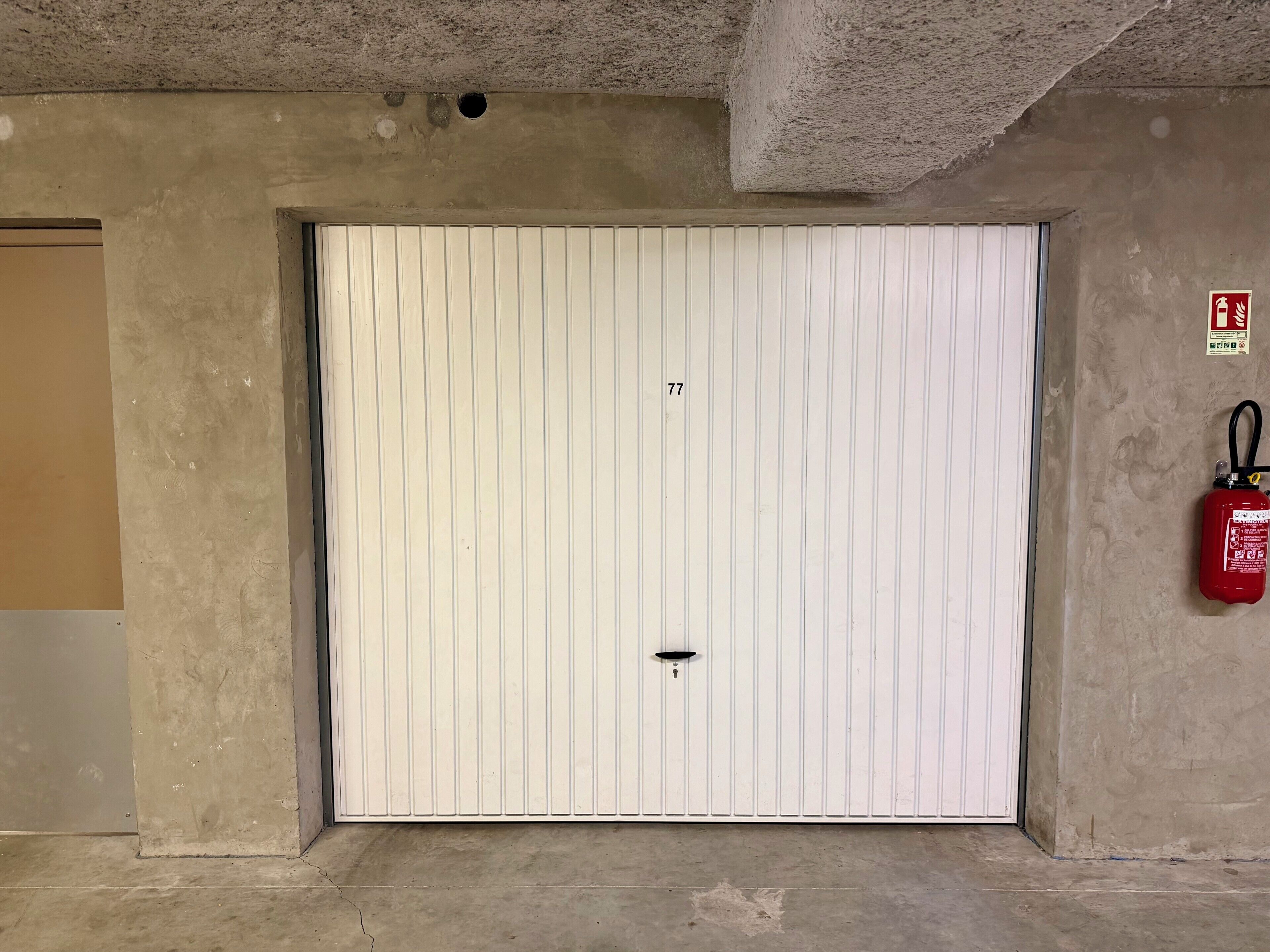 Une porte blanche portant le numéro « 77 », et un extincteur fixé au mur, à droite.
