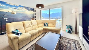 A living room with a sofa, a coffee table, a lamp, a TV, and a large window with a city and mountains view. - Comfortable T3 with terrace and garage! (Aix-les-Bains)