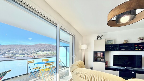 A balcony with a view of a body of water and distant hills, a sofa, a TV, and a lamp. - Comfortable T3 with terrace and garage! (Aix-les-Bains)
