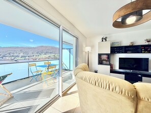 Ein Balkon mit Blick auf ein Gewässer und sanfte Hügel, eine Couch, ein Fernseher und eine Lampe.