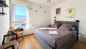 A bedroom with a bed, a chair, a window with a view, a radiator, and wall decorations. - Comfortable T3 with terrace and garage! (Aix-les-Bains)