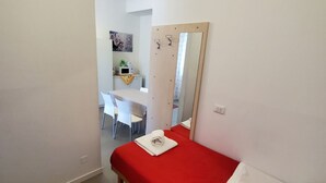 Una stanza piccola e pulita con una coperta rossa, una sedia bianca e una scrivania con uno specchio e una piccola pianta.