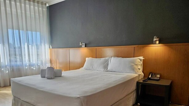 Une chambre d’hôtel moderne avec un grand lit, une tête de lit en bois, une table de chevet avec un téléphone et une fenêtre ornée de rideaux légers.