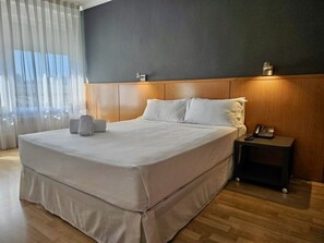 Ein modernes Hotelzimmer mit einem großen Bett, einem Holzkopfbrett, einem Nachttisch mit Telefon und einem Fenster mit durchsichtigen Vorhängen.