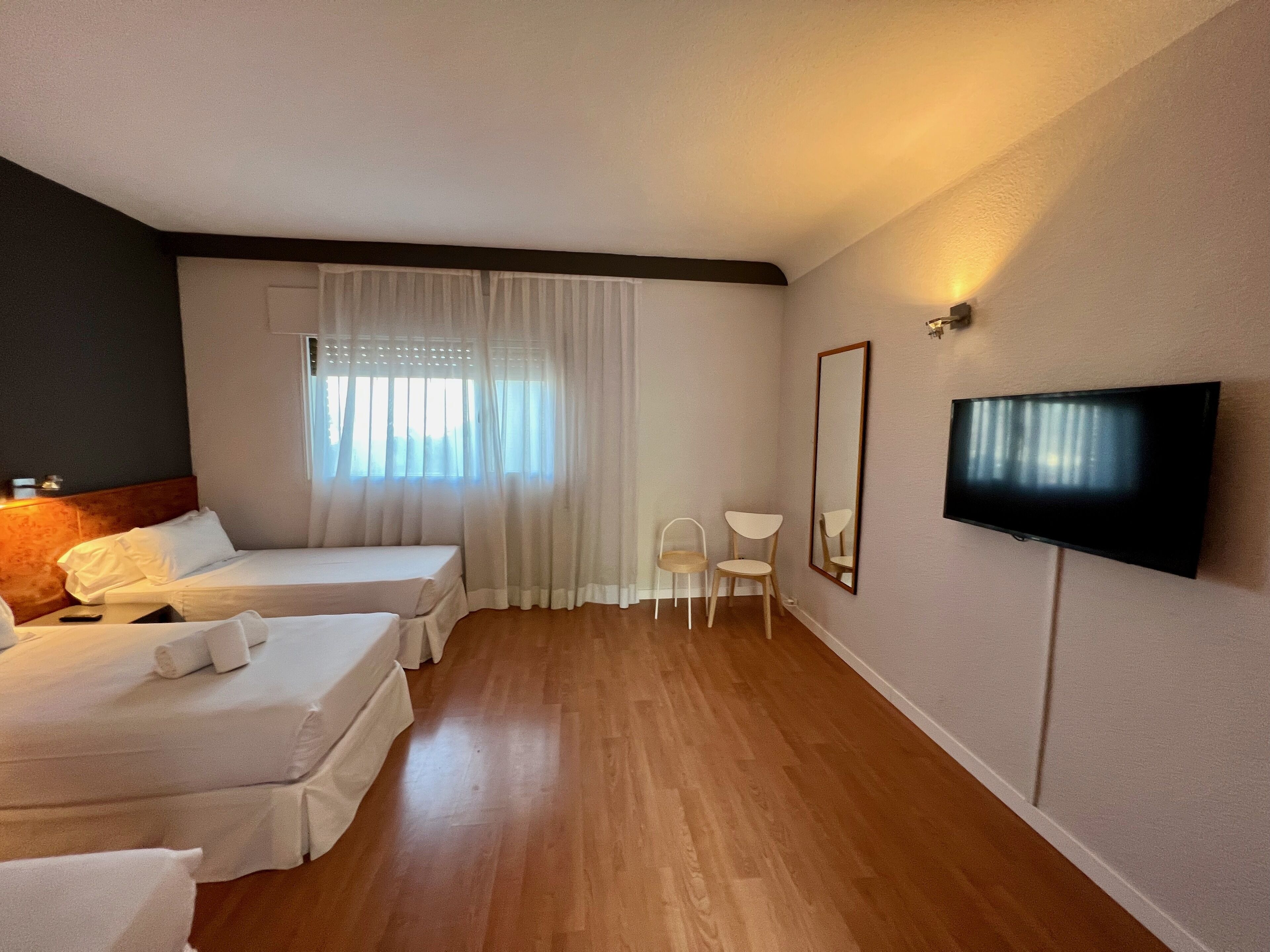 A hotel room with two beds, a TV, a small table, and a window with sheer curtains.