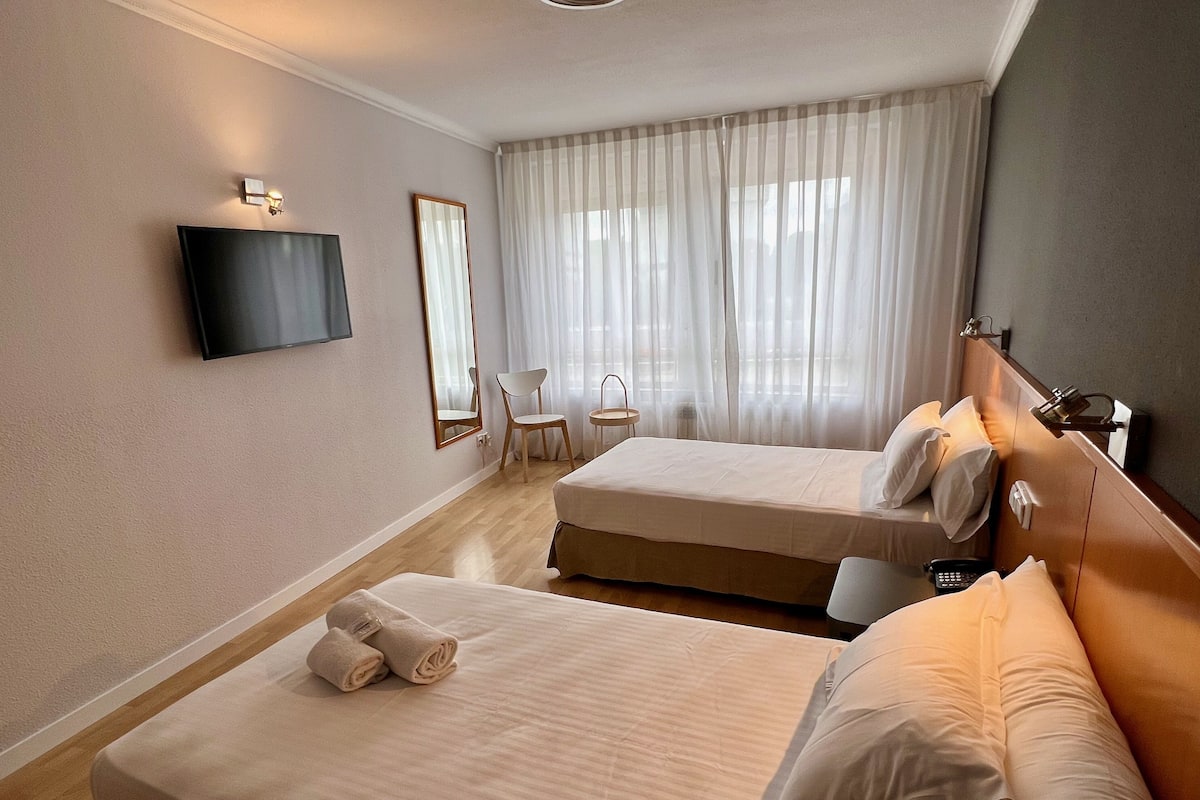 Ein Hotelzimmer mit zwei Betten, einem an der Wand montierten Fernseher, einem Sessel und einem Fenster mit durchsichtigen Vorhängen.
